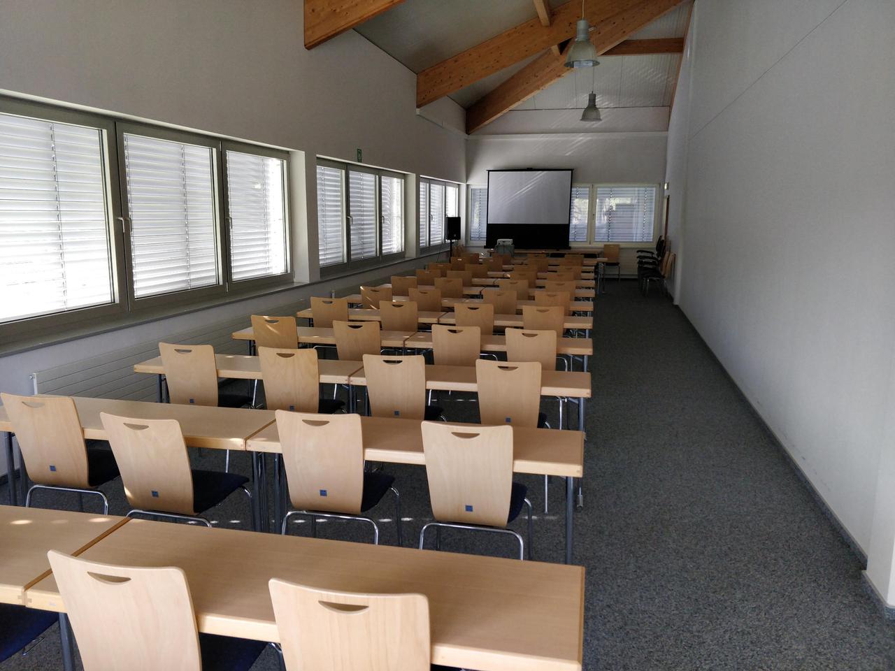 Bild eines sehr langen, schmalen Raumes. Es gibt 11 Stuhlreihen mit
jeweils nur 4 Stühlen pro Reihe, weil der Raum so schmal ist, dass
mehr nicht Platz haben. Das Foto ist von hinten aufgenommen. Vorne, in
weiter Ferne, ist eine Leinwand zu sehen, die vielleicht 1/6 der
Stirnfläche des Raumes ausfüllt, also für die Raumgröße zu klein
bemessen ist,